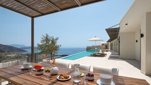 Una mesa de madera con platos, cuencos y vasos, con vistas a una piscina y al mar.