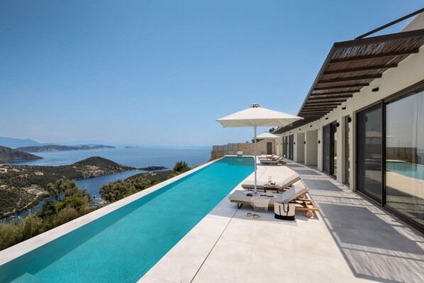 Uma piscina infinita moderna com vista desobstruída para o mar e colinas, área de estar externa com guarda-sóis e uma grande porta de vidro que dá acesso à piscina.