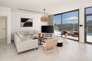 Uma sala de estar moderna com sofá, mesa de centro, televisão e cadeiras de vime. Apresenta uma grande janela com vista para uma piscina e montanhas.