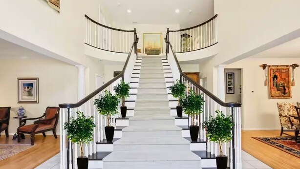 A grand staircase with a white railing, flanked by potted plants, leading to a landing with a framed picture.