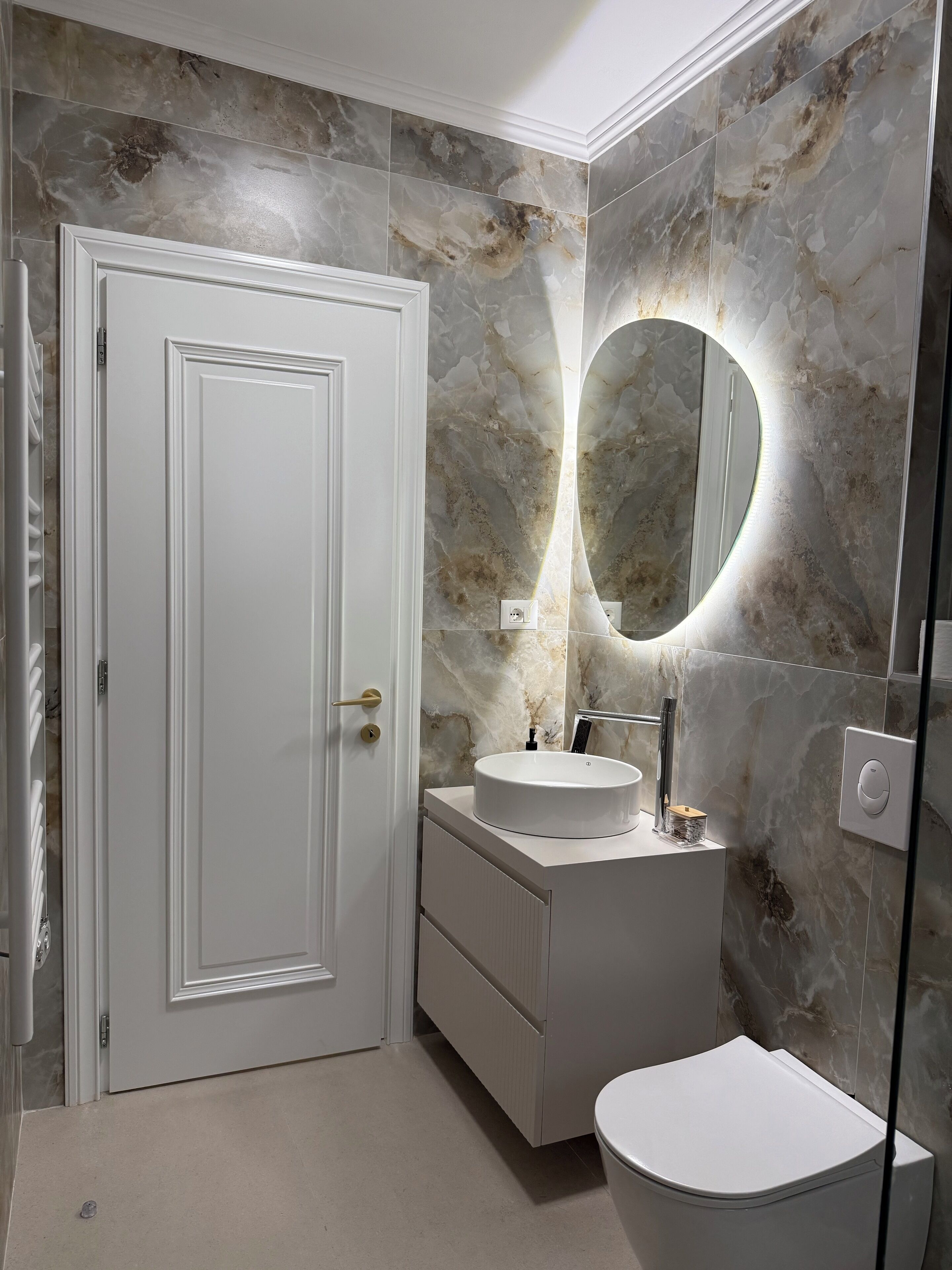 A bathroom with a white door, a round mirror with a light, a sink, and a toilet.