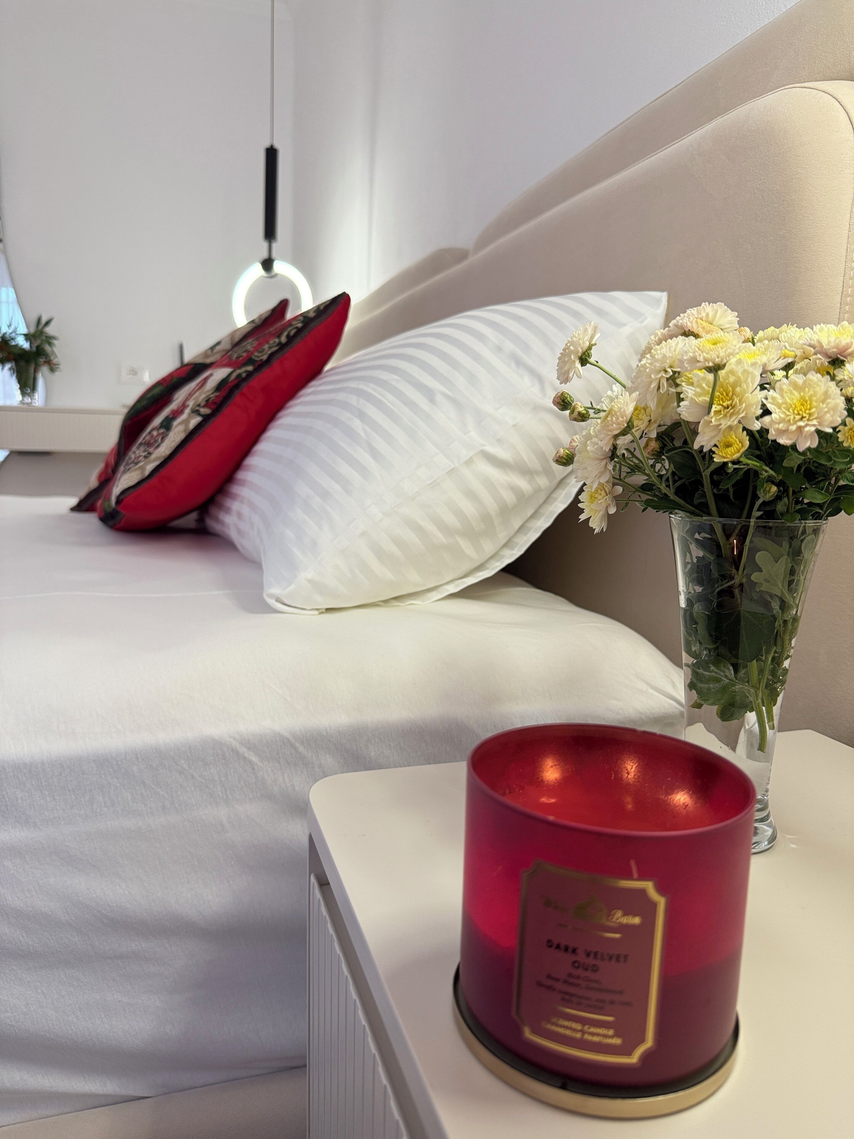 Un lit bien fait, avec du linge de lit blanc, un oreiller rouge, un vase de fleurs et une bougie rouge posée sur une table de chevet.