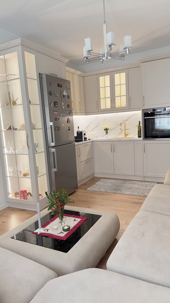 A modern kitchen with white cabinetry, a built-in refrigerator, a stove, and a dining area with a sofa and a coffee table.
