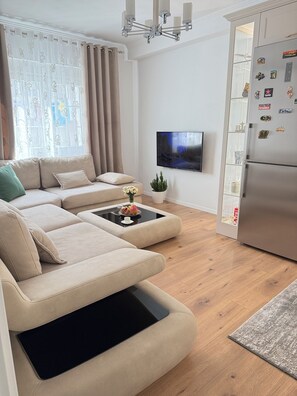 A modern living room with a sectional sofa, a coffee table, a flat-screen TV, a refrigerator, and decorative magnets.