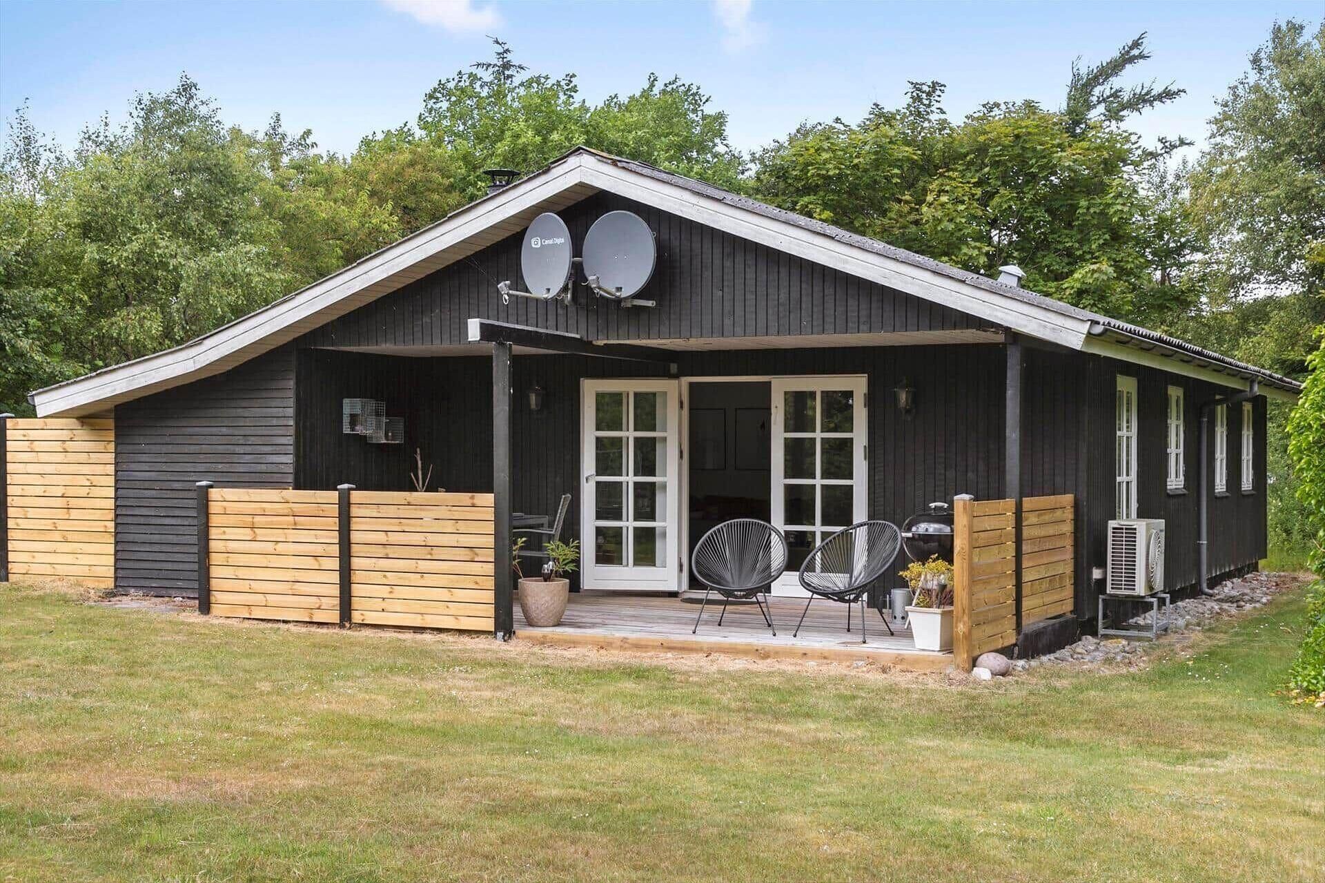 A black wooden building with a sloped roof, two satellite dishes, a covered porch with two chairs, and an air conditioning unit.