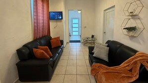 Een moderne woonkamer met twee zwarte leren banken, waarvan er een oranje kussens heeft, een glazen salontafel en een aan de muur bevestigde televisie.