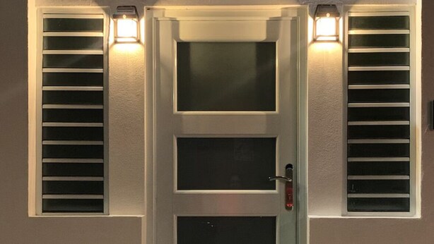 Een moderne deur met een ruitpaneel, met aan weerszijden twee verlichte wandlampen, en een kleine trap die er naartoe leidt.
