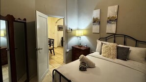 Een slaapkamer met een bed, een nachtkastje met lamp, een deur en een gang naar een eetgedeelte.