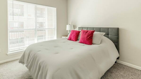 Een net opgemaakt bed met een grijs hoofdbord, wit beddengoed, twee rode kussens en een witte nachtlamp.