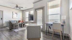 Una sala de estar moderna con un sofá gris, una mesa de centro redonda y un televisor. Hay ventanas amplias con persianas, un ventilador de techo y una puerta que da al exterior.