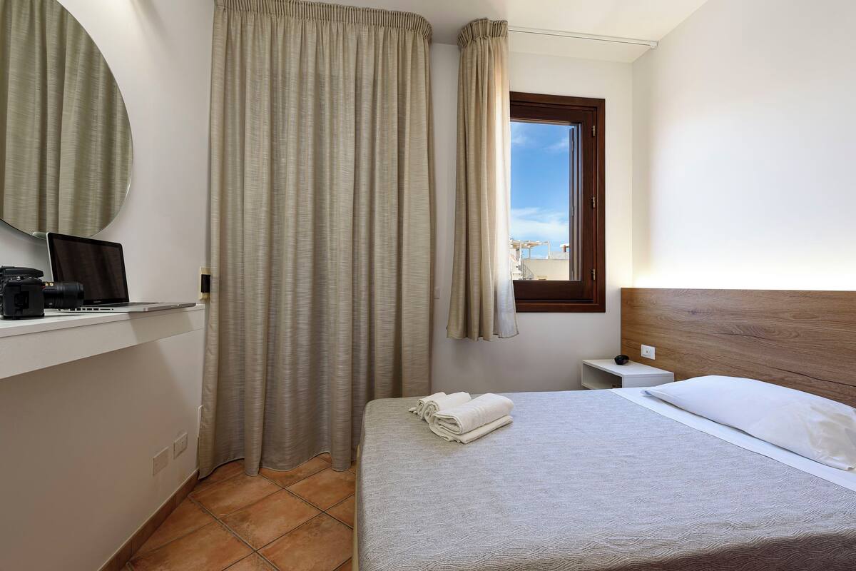 Ein Hotelzimmer mit einem Bett, einem Schreibtisch mit Laptop, einem Nachttisch und einem Fenster mit Vorhängen.