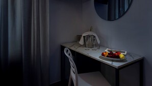 Een kleine, donkere kamer met een enkele witte stoel, een tafel met een schaal fruit en een spiegel aan de muur.