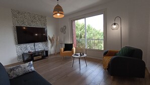 Un salon moderne doté d’un téléviseur à écran plat, d’un canapé, d’un fauteuil, d’une table basse et d’une grande fenêtre donnant sur les arbres.