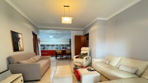 Ein modernes Wohnzimmer mit einer Couch, Hocker und einem weißen Couchtisch. Im Hintergrund befindet sich ein Essbereich mit Tisch und Stühlen.