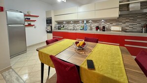 Eine moderne Küche mit Essbereich. Sie beinhaltet einen Kühlschrank, eine Mikrowelle, einen Ofen und eine Kaffeemaschine. Der Esstisch ist mit einer gelben Tischdecke und roten Stühlen gedeckt.