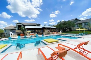 A swimming pool with lounge chairs and umbrellas, surrounded by buildings with stone and metal roofing. - Landing | Stylish 2BD, Pool, Gym (San Antonio)