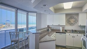 Une cuisine moderne avec des meubles blancs, des plans de travail en granit et une vue sur la plage grâce à de grandes fenêtres.