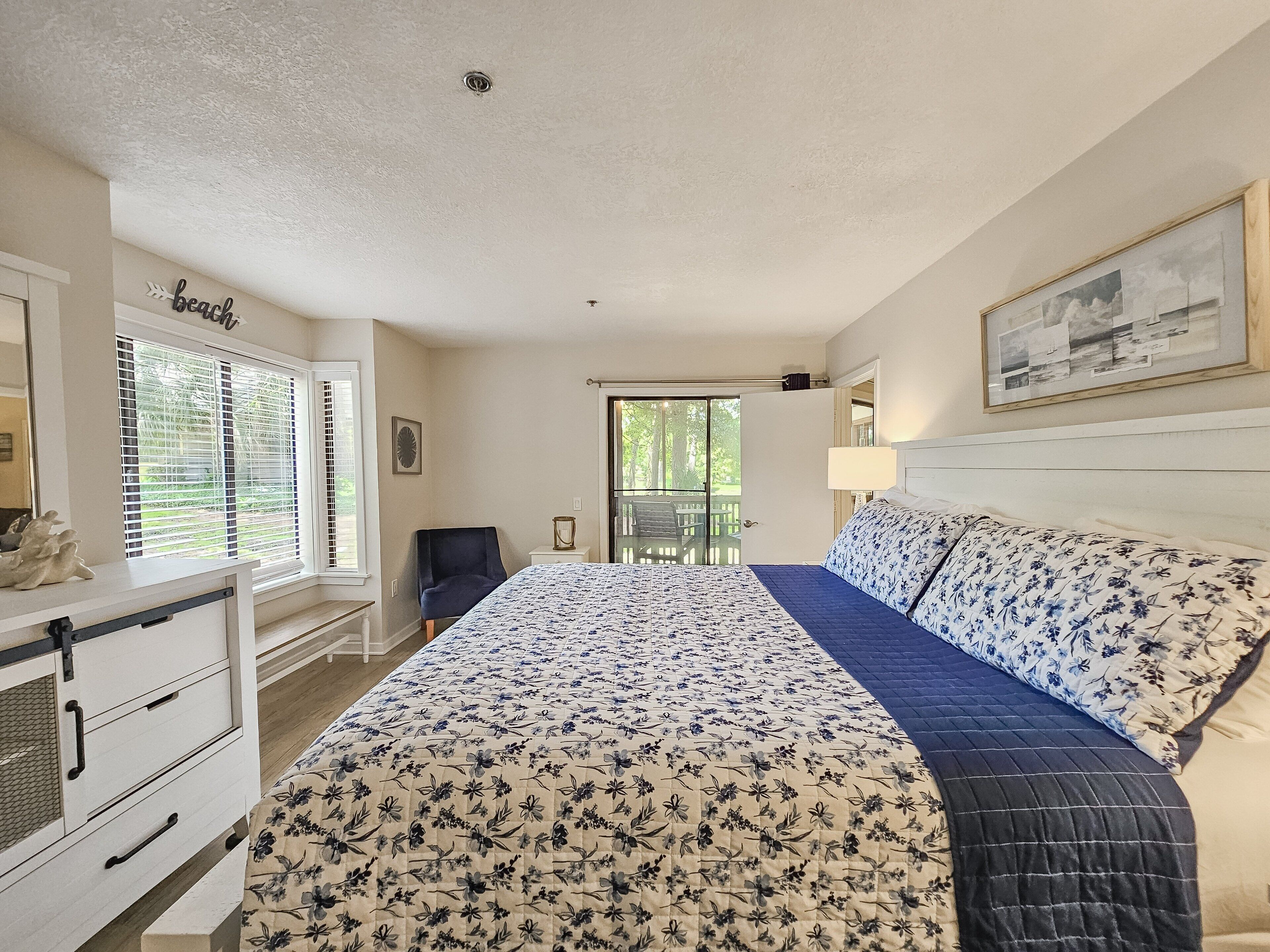 A bedroom with a large bed, a desk, and a chair. There is a window with blinds, a door leading to an outdoor area, and a painting on the wall.