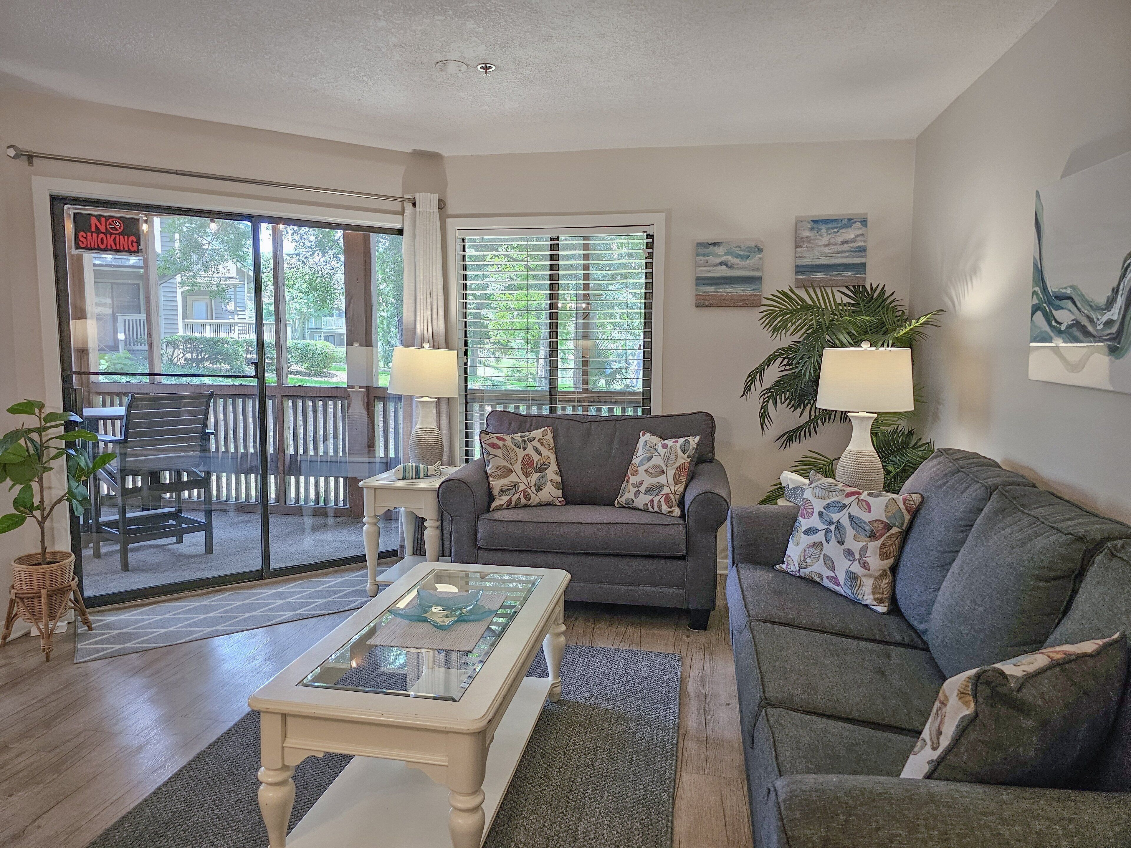 A living room with a grey sofa, a white coffee table, and a glass-top table. There are two lamps on side tables, a potted plant, and a view of the outside through a sliding glass door.