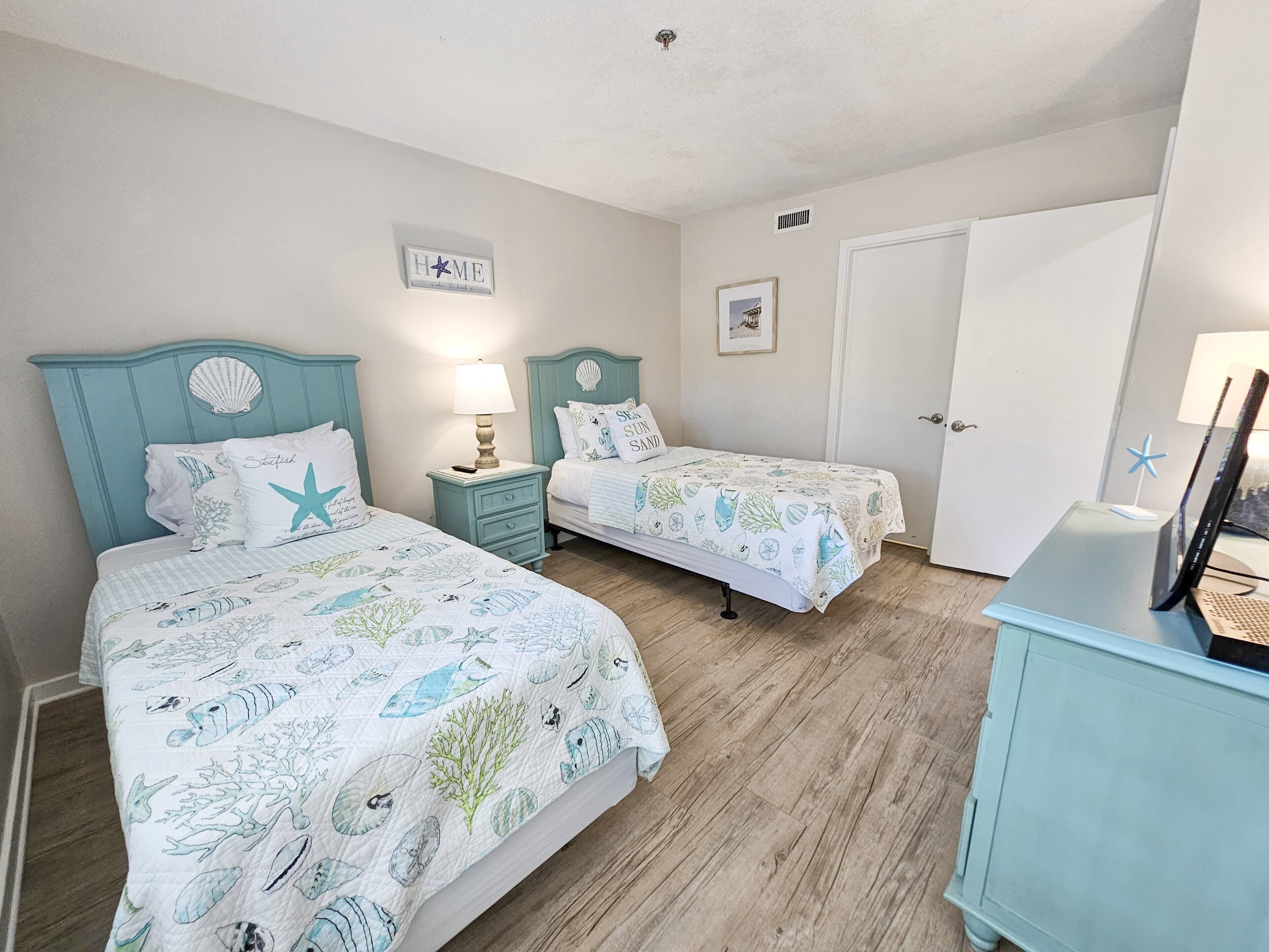 A bedroom with two beds, a nightstand with a lamp, a framed picture on the wall, and a 'HOME' sign.