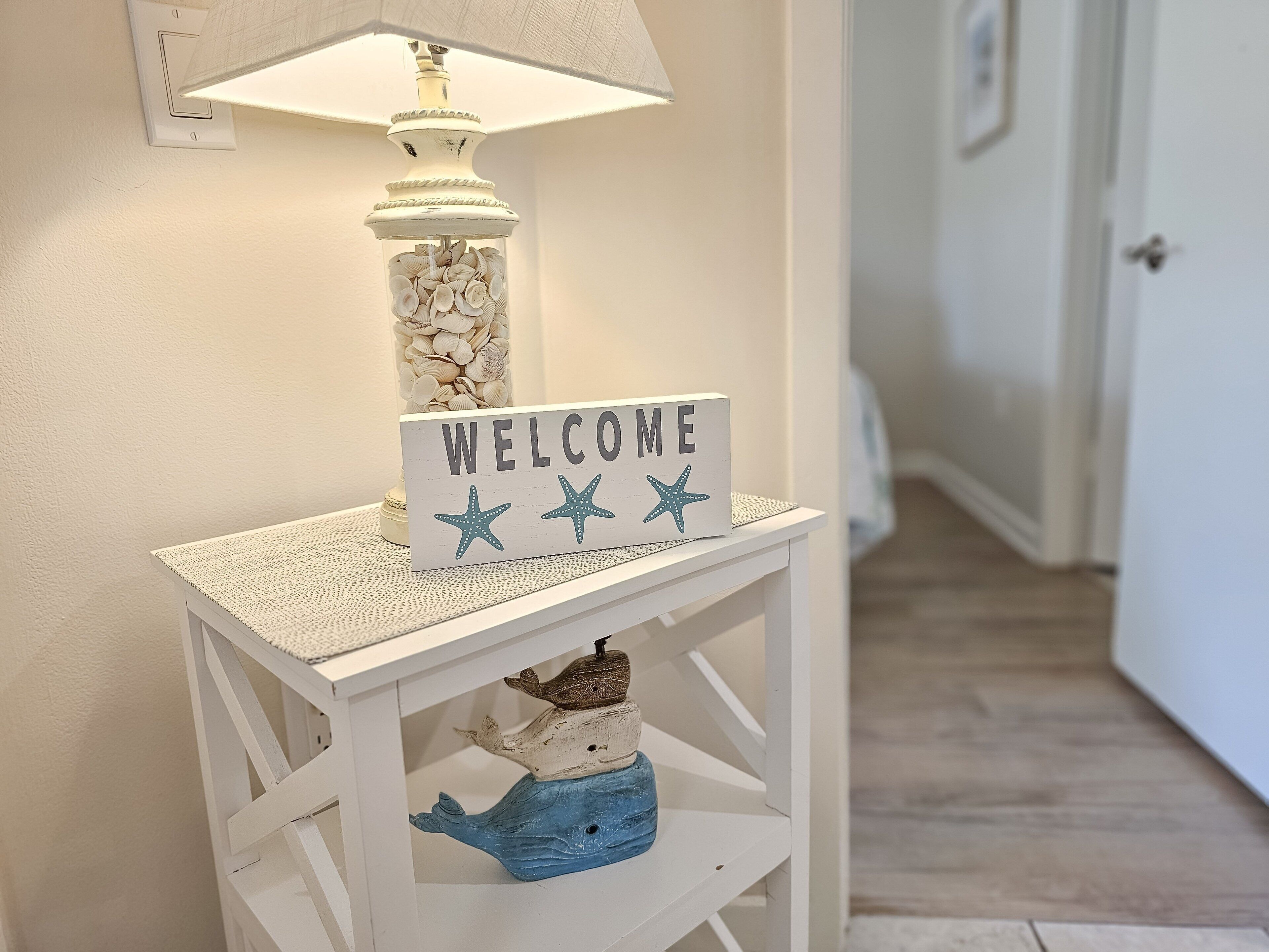 Una consola blanca con un cartel que dice 'WELCOME', una lámpara decorativa y una repisa con una figurita de ballena.
