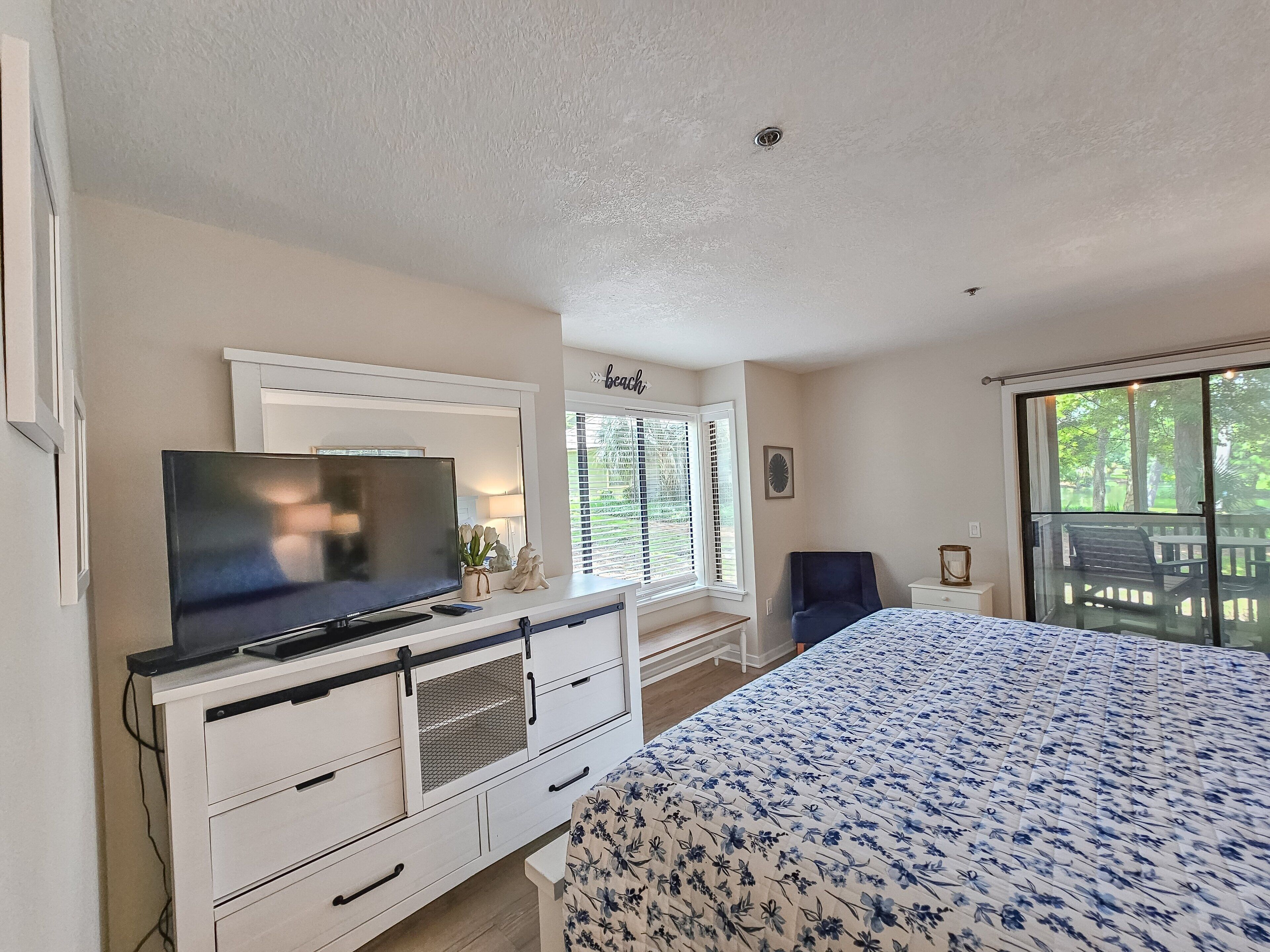 A bedroom with a bed, a TV on a dresser, a window with blinds, and a view of the outdoors.