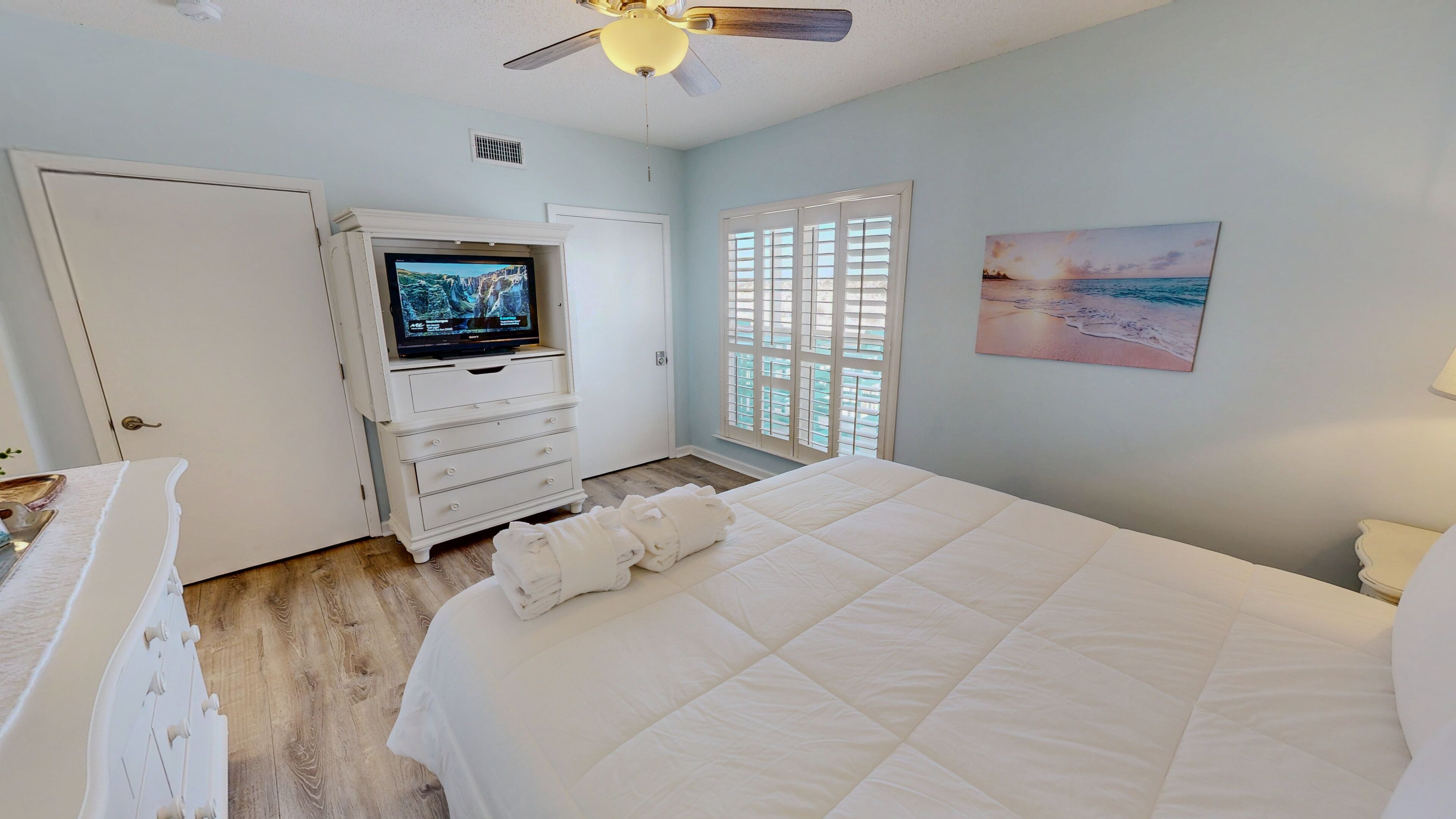 A bedroom with a bed, a TV on a dresser, a painting, and a window with shutters.