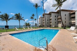 A swimming pool with lounge chairs and palm trees, surrounded by a building with balconies. - Maalaea Kai #102 1 Bedroom Condo (Wailuku)