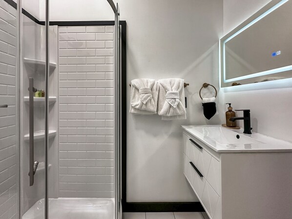 A modern bathroom with a glass shower enclosure, white tiled walls, a white sink with a black faucet, and a mirror above the sink.