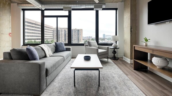 A modern living room with a grey sofa, a coffee table, and a TV on the wall. - Landing | Stunning 1BD, Game Room, Gym (Falls Church)