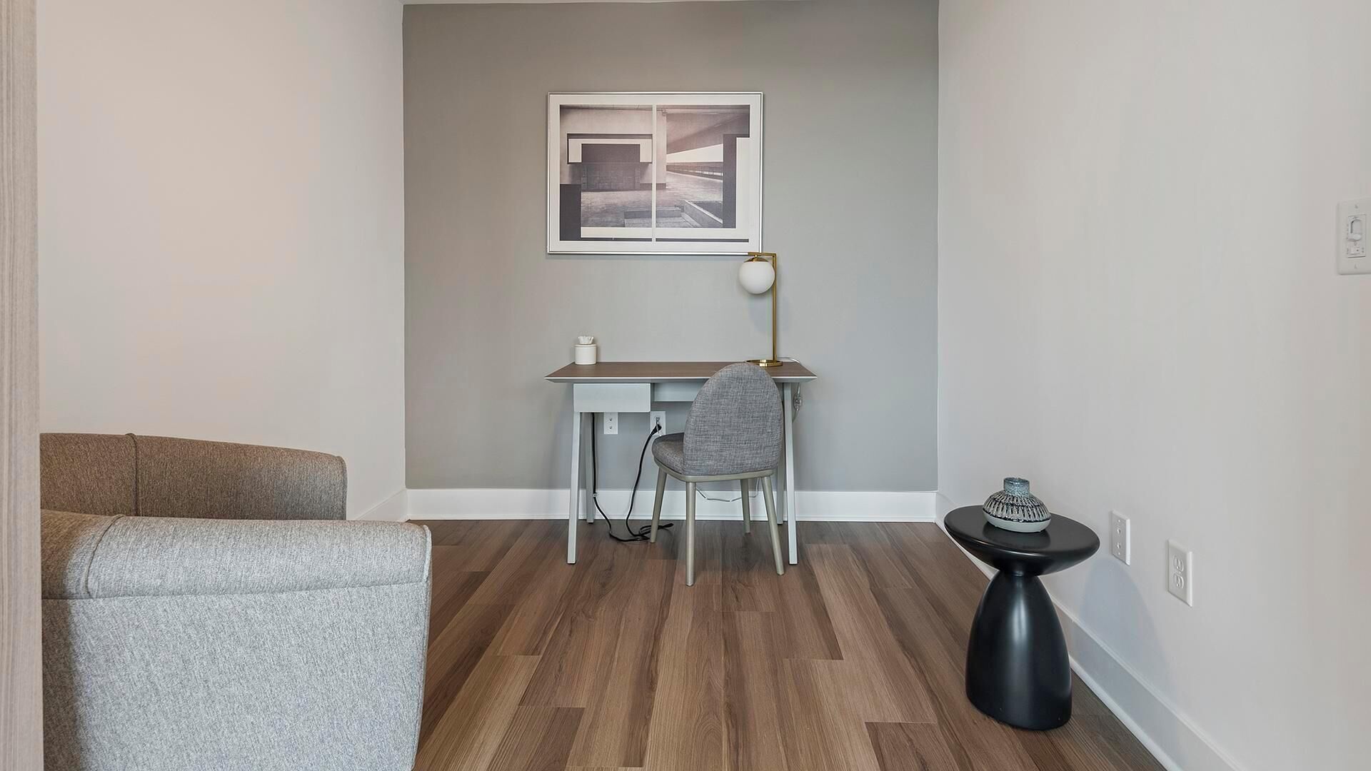 A modern living room with a grey sofa, a desk with a chair, a floor lamp, and a small side table with a decorative item.
