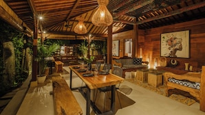 A spacious room with a wooden dining table, chairs, a sofa, and a bench. There are hanging light fixtures and a large painting on the wall. - Villa Dharuna Ubud (Ubud)
