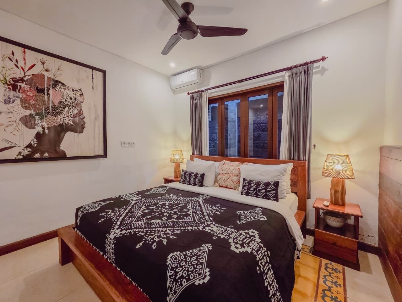 Ein Schlafzimmer mit Bett, Deckenventilator, Klimaanlage, Fenster mit Vorhängen und Nachttisch mit Lampe.