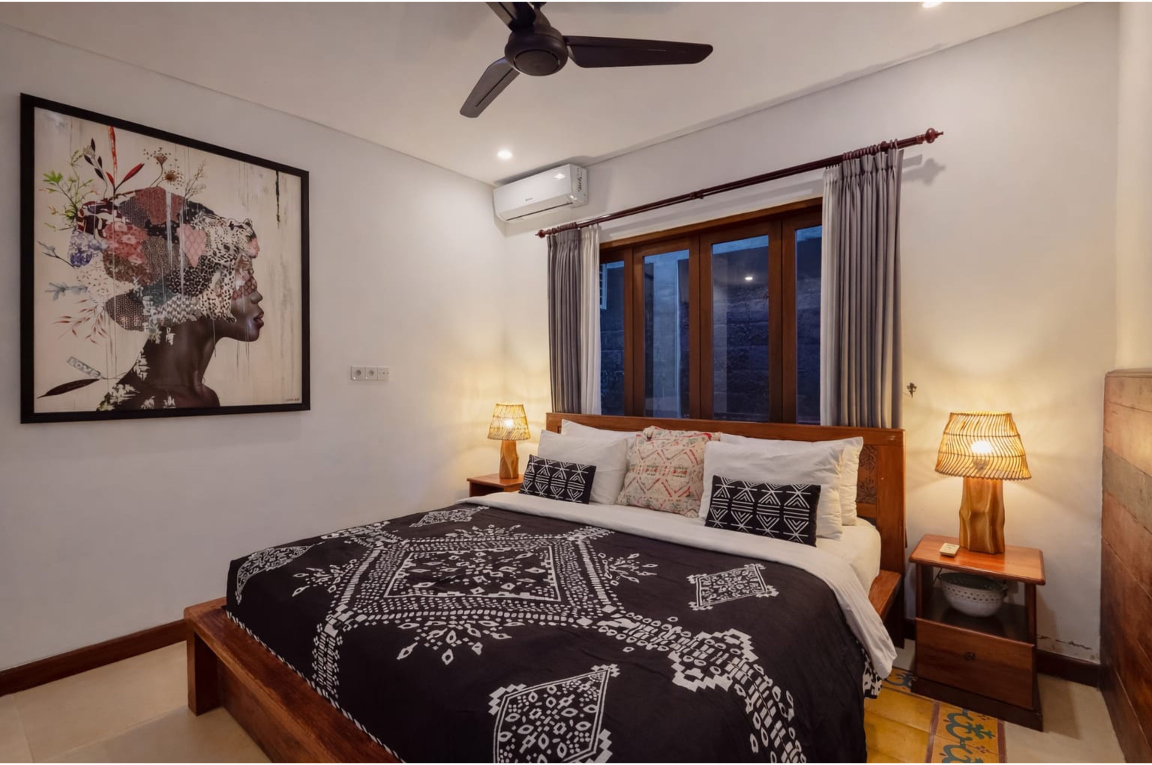 A bedroom with a bed, two bedside lamps, a ceiling fan, an air conditioning unit, a window with curtains, and a wall-mounted artwork.