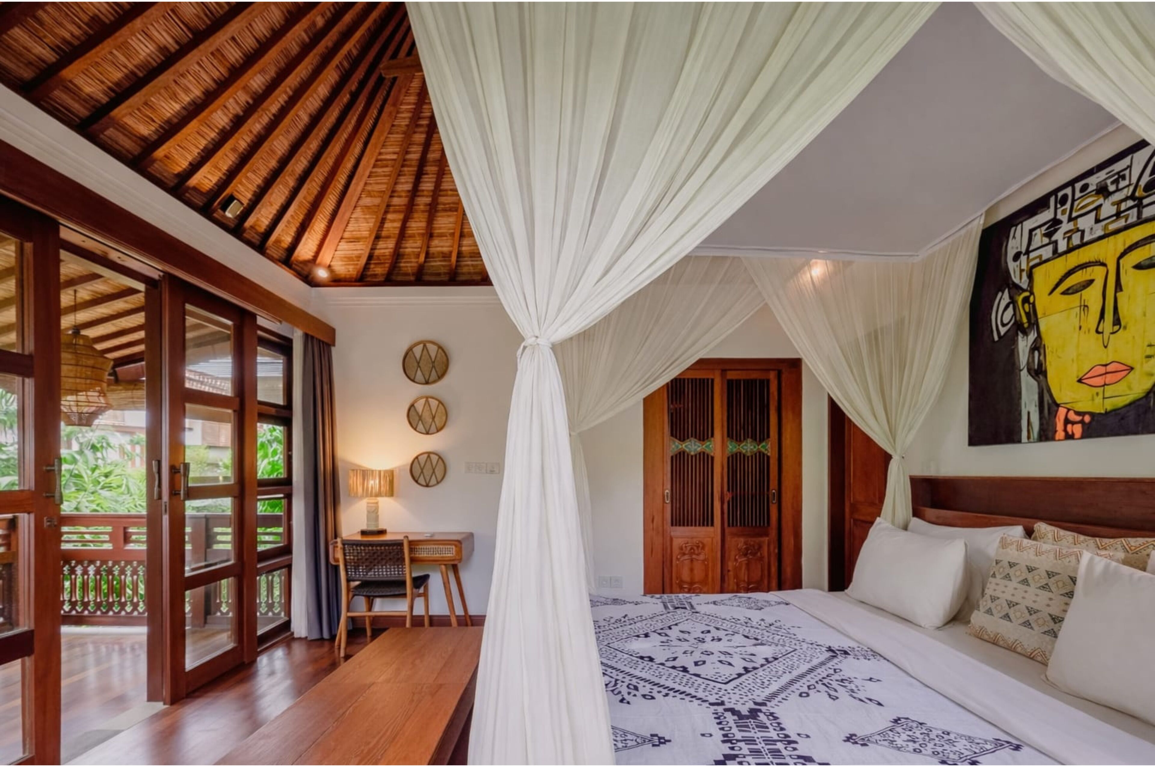 Quarto com cama, escrivaninha e cadeira, uma janela grande e porta que dá para área externa.