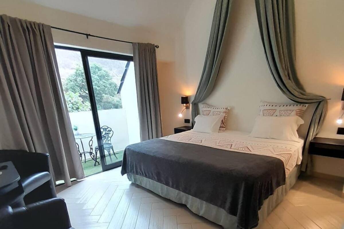 A bedroom with a large bed, a balcony with chairs, and a view of greenery outside.