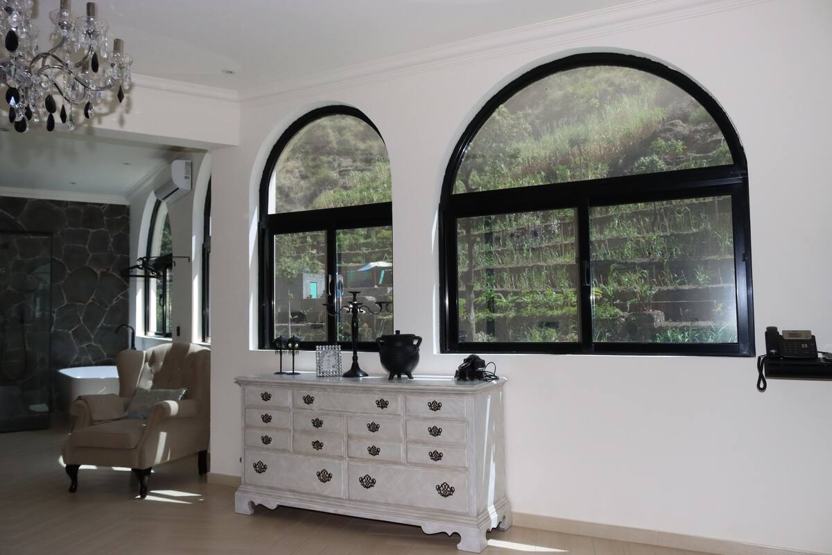 A spacious living room with a large arched window, a white dresser, a beige armchair, and a chandelier.