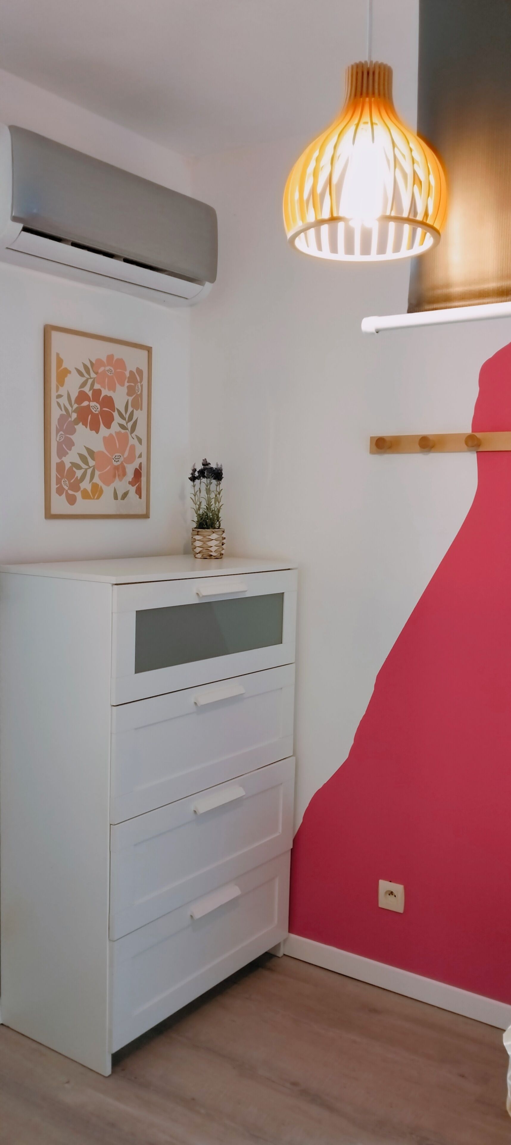 A room with a white dresser, a wall-mounted air conditioner, a hanging light fixture, and a red accent wall.