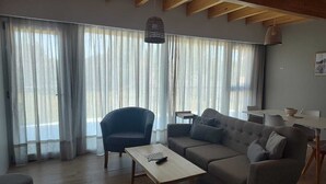 A living room with a sofa, armchair, and a small table. There are curtains on the windows, and a ceiling with wooden beams.