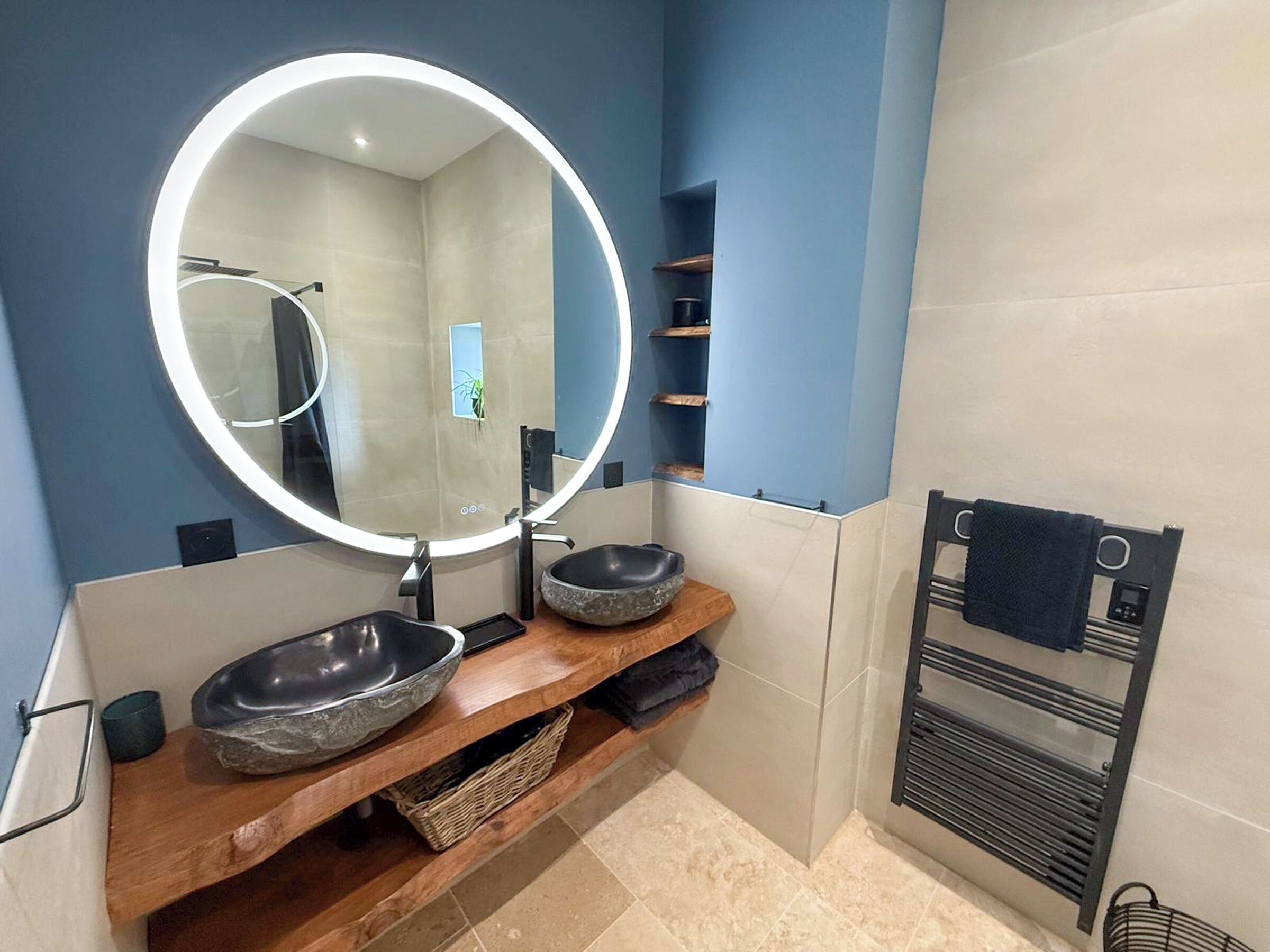 A bathroom with a round mirror, two stone bowls, a wooden vanity, a towel rack, and a wall-mounted heater.