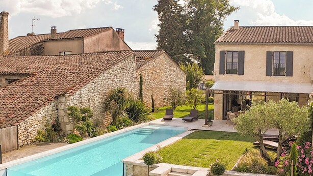 A swimming pool surrounded by a well-maintained garden with a stone wall, a grassy lawn, and a building with a tiled roof.