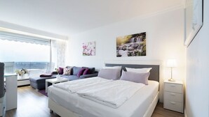 A modern bedroom with a large bed, a sofa, a small table, and a nightstand. There is a window with a view of the sea, and a painting on the wall. - Suite auf Wolke 7 von OstseeAhoi (Timmendorfer Strand)
