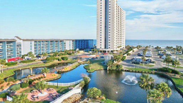 A high-rise hotel with a pool area, including a fountain, surrounded by greenery and a parking lot.