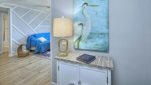 A room with a blue bed, a lamp, and a wall painting of a bird.
