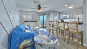 A modern living room with a blue sofa, a patterned ottoman, and a dining area with wooden chairs and a white table.