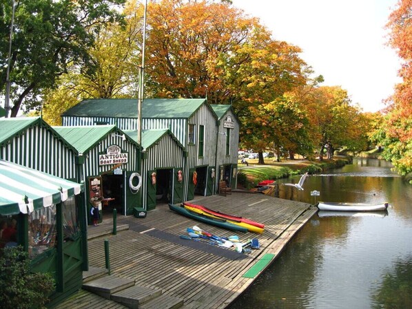 Un cobertizo para botes con techo verde y paredes a rayas, un muelle con canoas y un pequeño bote en el agua.