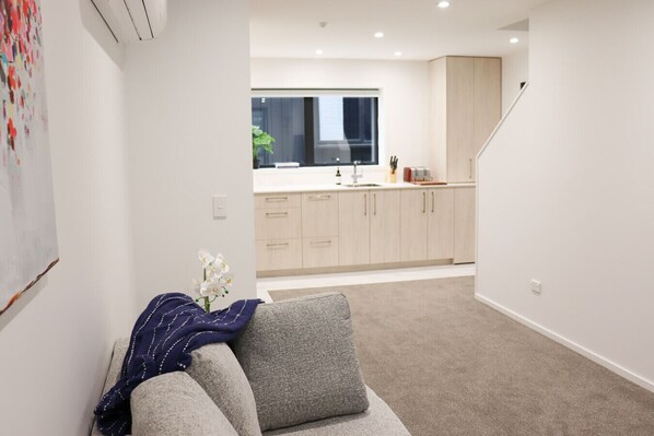 Un apartamento moderno con un sofá gris, una mesita con un jarrón de flores y una cocina con armarios de madera clara y una ventana.
