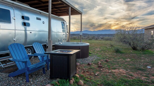 Ein Wohnmobil mit zwei blauen Adirondack-Stühlen, einem Whirlpool und einem Grill vor einem überdachten Bereich mit Blick auf Berge und einen Sonnenuntergang.
