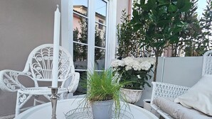 Una mesa redonda blanca con una vela y una planta en maceta, rodeada de mobiliario de mimbre blanco y un banco acolchado blanco.