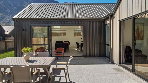 A modern outdoor patio with a wooden table and chairs, a glass door leading to an interior space with a sofa and chairs, and a mountainous backdrop.